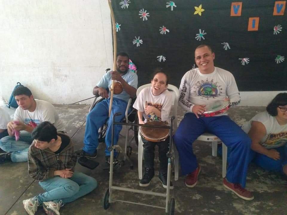 Descrição da imagem #PraCegoVer: Imagem no formato retangular, na horizontal. Um grupo de pessoas com deficiência tocando instrumentos durante uma roda de capoeira. Fim da descrição.