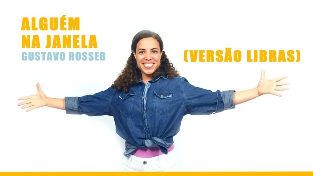Descrição da imagem #PraCegover: Imagem no formato retangular, na horizontal. Cena do clipe em Libras. Na parte central da imagem está uma mulher morena, de cabelos castanhos, cacheados, abaixo dos ombros. Ela usa uma camisa jeans e está com os braços abertos. Na lateral esquerda está escrito, em letras amarelas: Alguém na janela. E, em azul, abaixo, está o nome Gustavo Rosseb. Na lateral direita está escrito, em amarelo: versão em Libras. Fim da descrição.