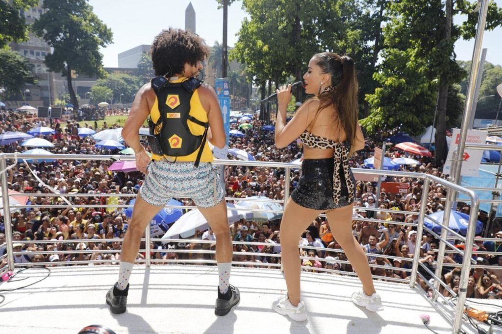 Descrição da imagem #PraCegoVer: Imagem no formato retangular, na horizontal. A cantora Anitta e o educador surdo Leo Castilho estão em cima do trio elétrico. Leo está usando a mochila especial. Eles estão dançando, lado a lado. Fim da descrição.