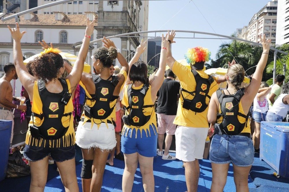Descrição da imagem #PraCegoVer: Imagem no formato retangular, na horizontal. Grupo de pessoas surdas convidadas para testar a mochila. Elas estão em cima do trio elétrico e usam uma camiseta amarela e a mochila especial. Fim da descrição.