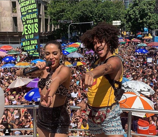 Descrição da imagem #PraCegoVer: Imagem no formato retangular, na horizontal. A cantora Anitta e o educador surdo Leo Castilho estão em cima do trio elétrico. Leo está usando a mochila especial. Eles estão dançando, lado a lado. Fim da descrição.