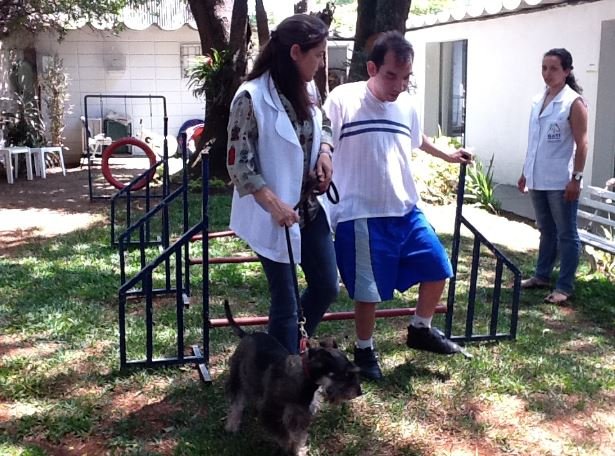 Descrição da imagem: Foto de um rapaz com deficiência. Ela está caminhando ao lado de uma mulher e de um cachorro. Fim da descrição.