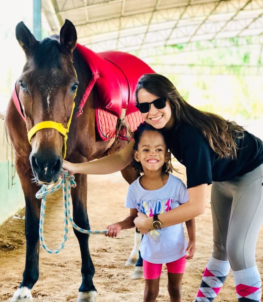 Descrição da imagem #PraCegoVer: Imagem no formato retangular, na vertical. Equoterapeuta posa com paciente ao lado do cavalo. Elas estão sorrindo. Fim da descrição.