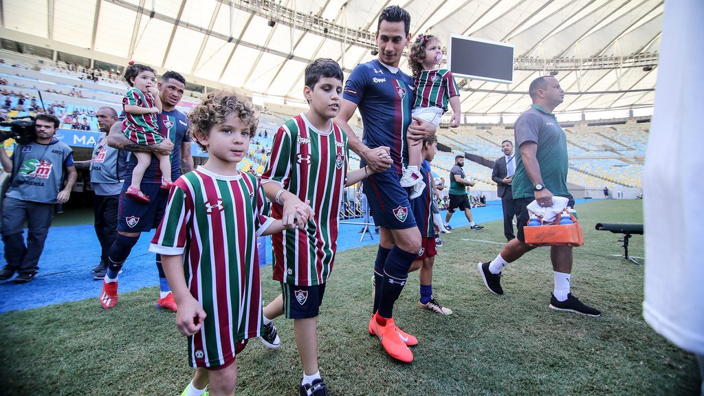 Descrição da imagem #PraCegoVer: Imagem no formato retangular, na horizontal. O menino Bernardo entrando em campo de mãos dadas com o jogador Ganso. Ao lado deles está outro menino, que assim como Bernardo, veste o uniforme do Fluminense. O jogador Ganso está com uma criança em seu colo. Ao fundo vemos o estádio do Maracanã. Fim da descrição.