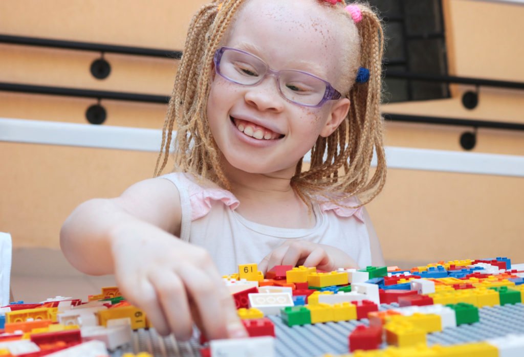 Descrição de imagem #pracegover: Foto de uma menina brincando com os blocos de montar em braille. A menina é albina, ela usa óculos e está sorrindo. Fim da descrição.