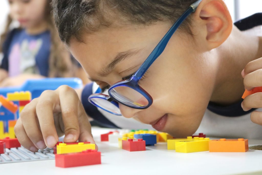 Descrição de imagem #PraCegoVer: Imagem no formato retangular, na horizontal. Um menino está brincando com os blocos de montar em braille. O menino usa óculos e está olhando as peças bem de perto. Fim da descrição.