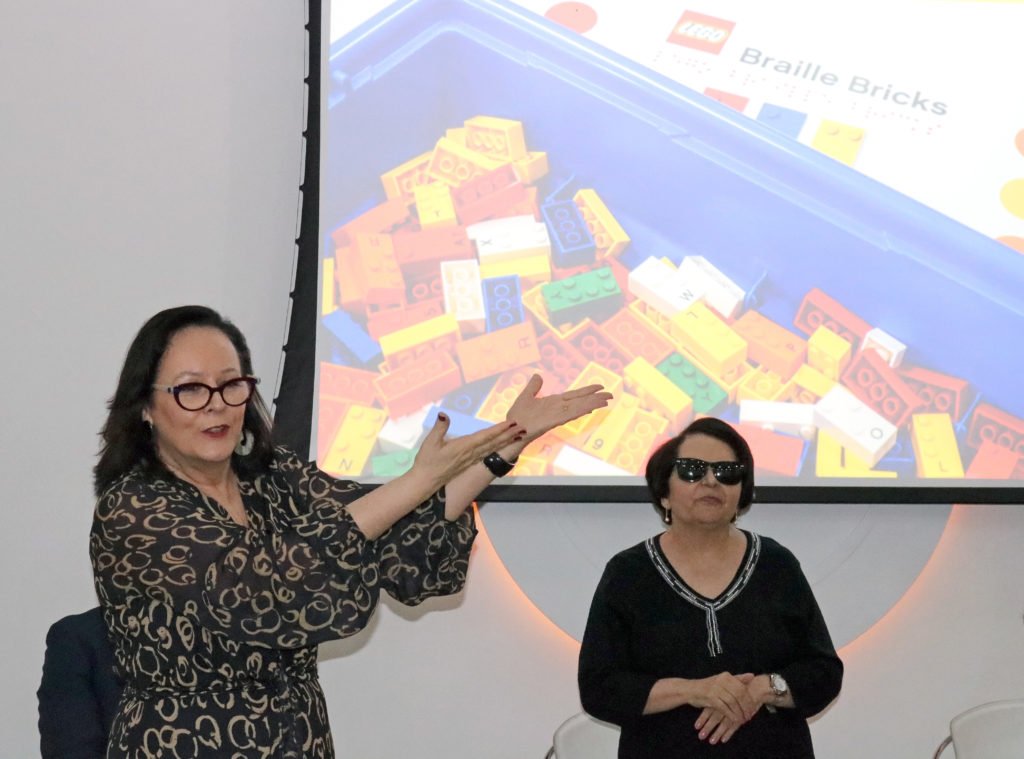 Descrição da imagem #PraCegoVer: Imagem no formato retangular, na horizontal. Duas mulheres estão à frente de um telão, que traz imagens dos blocos de montar em braille. Fim da descrição