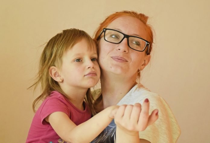 Descrição da imagem #PraCegoVer: Imagem no formato retangular, na horizontal. Uma mulher e uma menina representam a figura de mãe e filha. Elas estão abraçadas. A mulher é ruiva, tem a pele branca e usa óculos e uma camisa branca. A menina é loira, tem a pele clara e está usando uma blusa cor de rosa. Fim da descrição.