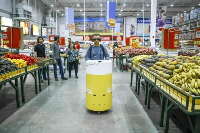 Descrição da imagem #PraCegoVer: Imagem no formato retangular, na horizontal. Um homem cego conduz o carrinho de compras acessível da Skol em um supermercado. O equipamento tem formato semelhante ao de uma lata de cerveja, está pintado de amarelo e branco, e tem a logo da Skol. Fim da descrição.