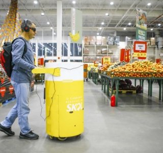 Descrição da imagem #PraCegoVer: Imagem no formato retangular, na horizontal. Um homem cego conduz o carrinho de compras acessível da Skol em um supermercado. O equipamento tem formato semelhante ao de uma lata de cerveja, está pintado de amarelo e branco, e tem a logo da Skol. Fim da descrição.
