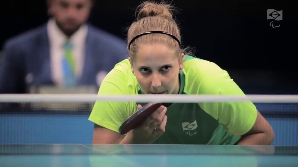 Descrição da imagem #PraCegoVer: Imagem no formato retangular, na horizontal. Uma atleta está disputando um torneio de tênis de mesa. Ela está abaixada, durante uma jogada. A atleta é loira e tem os cabelos lisos, que estão presos em um coque. Ela usa o uniforme verde e amarelo do CPB. Fim da descrição.