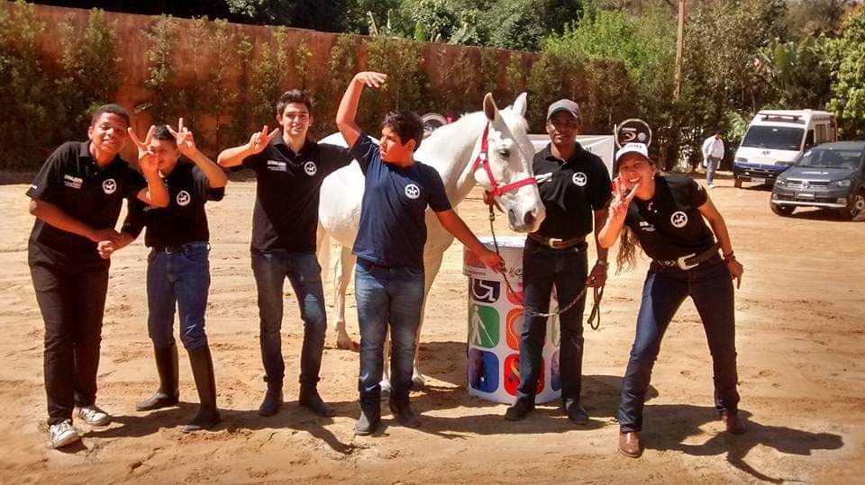 Descrição da imagem #PraCegoVer: Imagem no formato retangular, na horizontal. Equipe da Hípica Santa Terezinha durante prova dos 3 Tambores. Eles estão com Murilo. Eles estão na frende de um cavalo branco. Fim da descrição.