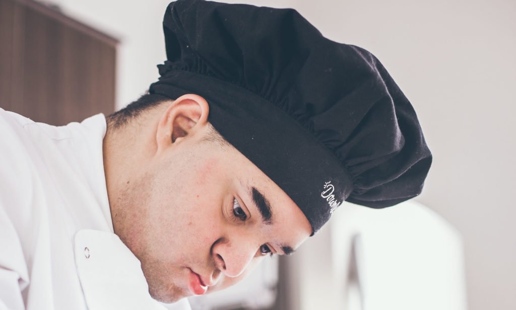 Descrição de imagem #PraCegoVer: Imagem no formato retangular, na vertical. Gabriel é um rapaz que tem síndrome de Down. Ele está preparando uma receita. Gabriel é moreno e tem a pele clara.. Ele usa uma Doma branca e um chapéu preto de cozinheiro,. Fim da descrição.