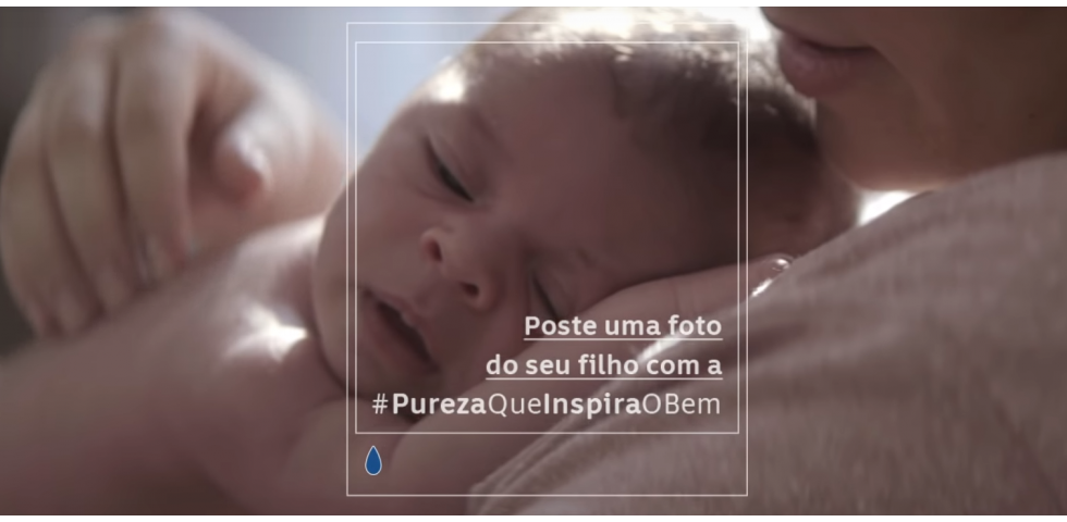 Descrição da imagem #PraCegoVer: Imagem no formato retangular, na horizontal. Um bebê está deitado no peito da mãe, que acaricia suas costas. No meio da imagem existem duas bordas quadradas, onde está escrito com letras brancas: Poste uma foto do seu filho com a #purezaqueinspiraobem. Fim da descrição.