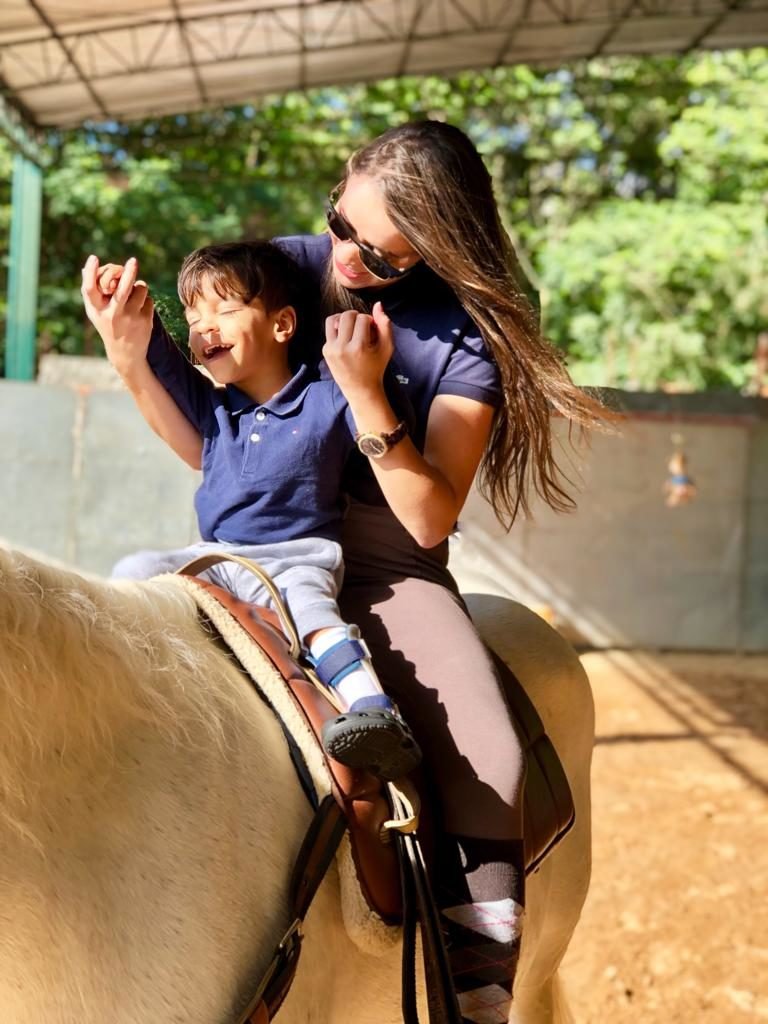 Descrição da imagem #PraCegoVer: Imagem no formato retangular, na vertical. Uma criança e uma equoterapeuta estão montados no cavalo. Fim da descrição.