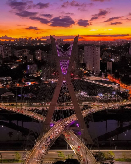 Descrição da imagem #PraCegoVer: Ponte Estaiada iluminada com a cor lilás. Ao fundo vemos um por do sol. Fim da descrição.