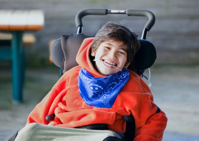 Descrição da imagem #PraCegoVer: Um menino com paralisia cerebral está sorrindo. Ele é moreno, tem pele clara e cabelos castanhos. O menino veste uma blusa de moletom vermelha, com capuz, e um lenço azul. Fim da descrição.