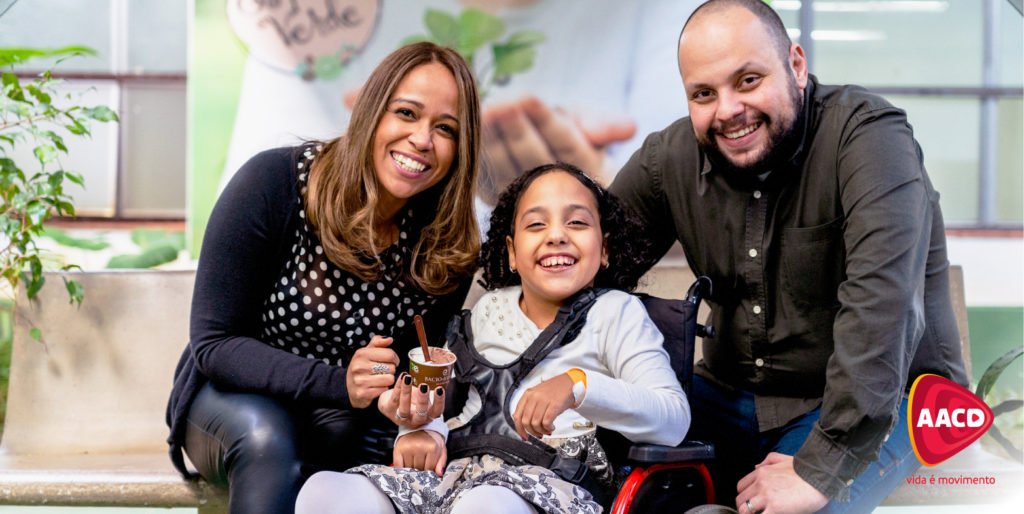 Descrição da imagem #PraCegoVer: Uma menina com deficiência física está em sua cadeira de rodas e seus pais estão ao seu lado. Eles estão sorrindo. No canto inferior da imagem está o logo da AACD. Fim da descrição.