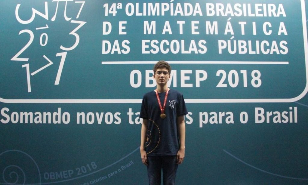Descrição da imagem #PraCegoVer: O jovem Gabriel, posa com sua medalha de ouro. Gabriel é um rapaz moreno, de pele clara e cabelos castanhos, curtos. Ele está de pé, no palco da premiação. Gabriel usa uma camiseta azul marinho e calças pretas. Ele também usa óculos de grau. Fim da descrição.