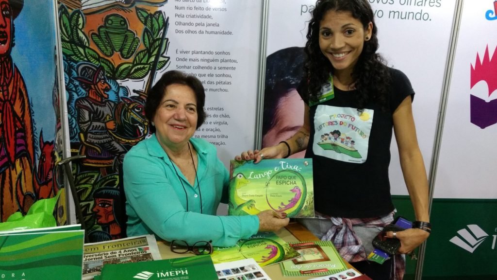 Descrição da imagem #PraCegoVer: A escritora Dinorá Couto Cançado durante uma sessão de autógrafos de seu livro. Ela está sentada e tem uma moça em pé, ao seu lado, recebendo o livro das mãos da escritora. Dinorá é uma mulher morena, de pele clara e cabelos castanhos, curtos. Ela está vestindo uma camisa verde e sorri. A moça ao lado dela é morena, de pele clara, e cabelos castanhos cacheados. Ela veste uma camiseta preta com uma ilustração branca. Fim da descrição.