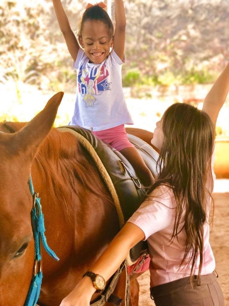 Descrição da imagem #PraCegoVer. Uma menina está montada em um cavalo, durante a sessão de equoterapia. Ela está acompanhada da equoterapeuta e está fazendo um movimento com os braços para cima. A menina é negra e está usando uma camiseta branca com desenhos coloridos e um shorts cor de rosa. A equoterapeuta é morena e está olhando para a menina. Elas estão no setting terapêutico. Fim da descrição.