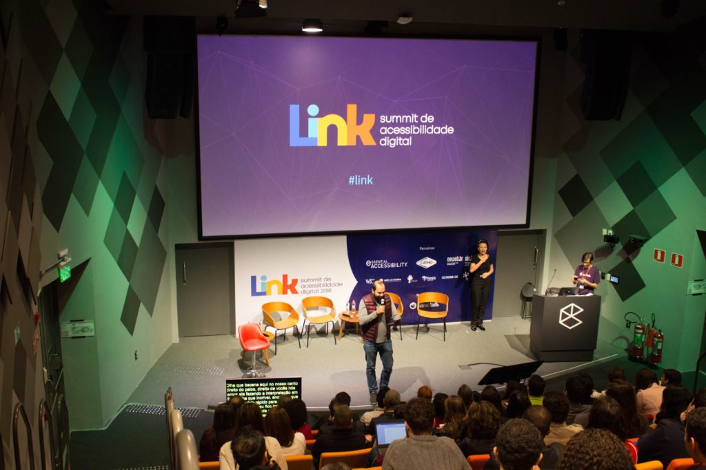 Descrição da imagem #PraCegoVer: O palco da primeira edição do evento da Hand Talk. Nele, está um palestrante e, ao fundo, uma intérprete de Libras. Fim da descrição.