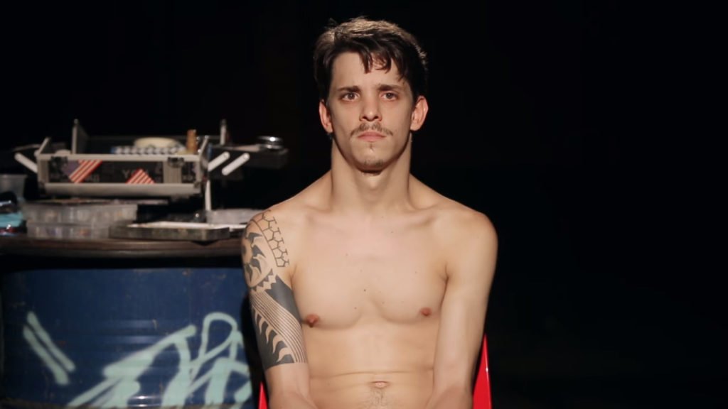 Descrição da imagem #PraCegoVer: Daniel está sentado em uma cadeira. Ele está sem camisa. Daniel é um rapaz moreno, de pele clara e cabelos castanhos, lisos e curtos. Fim da descrição.