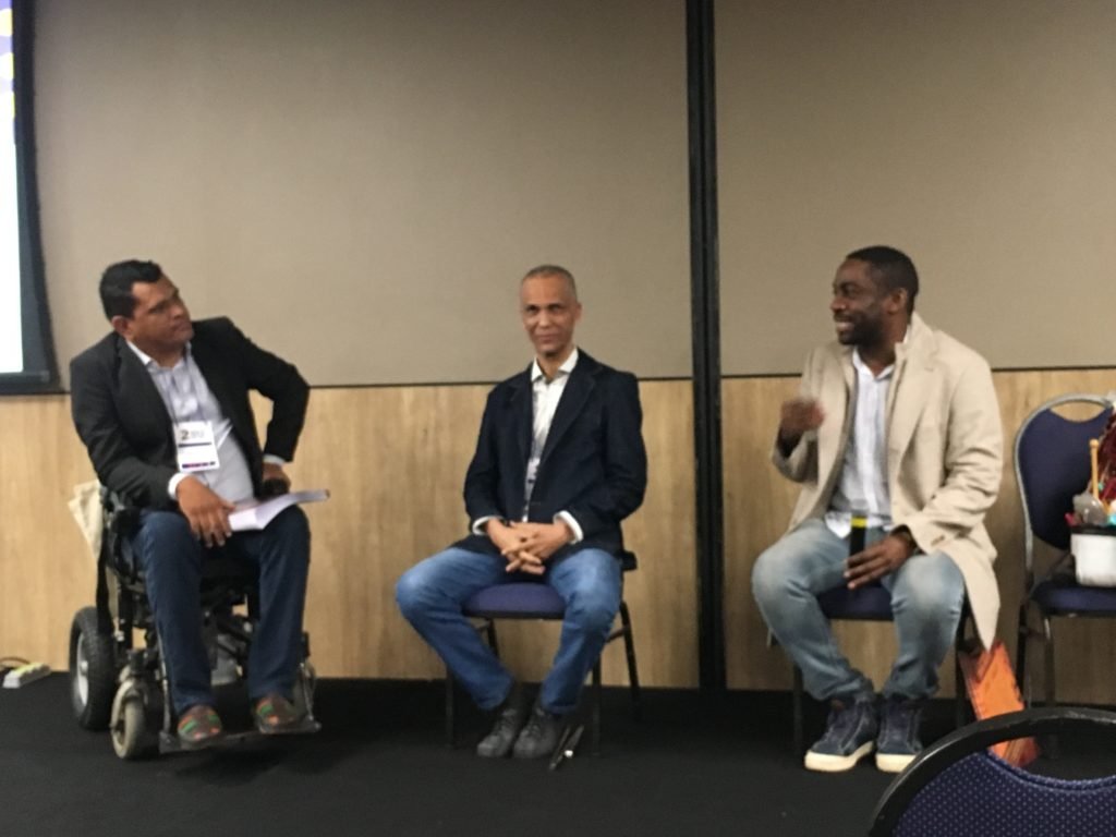 Descrição da imagem #PraCegoVer: Três homens estão sentados no palco do evento. Fim da descrição.
