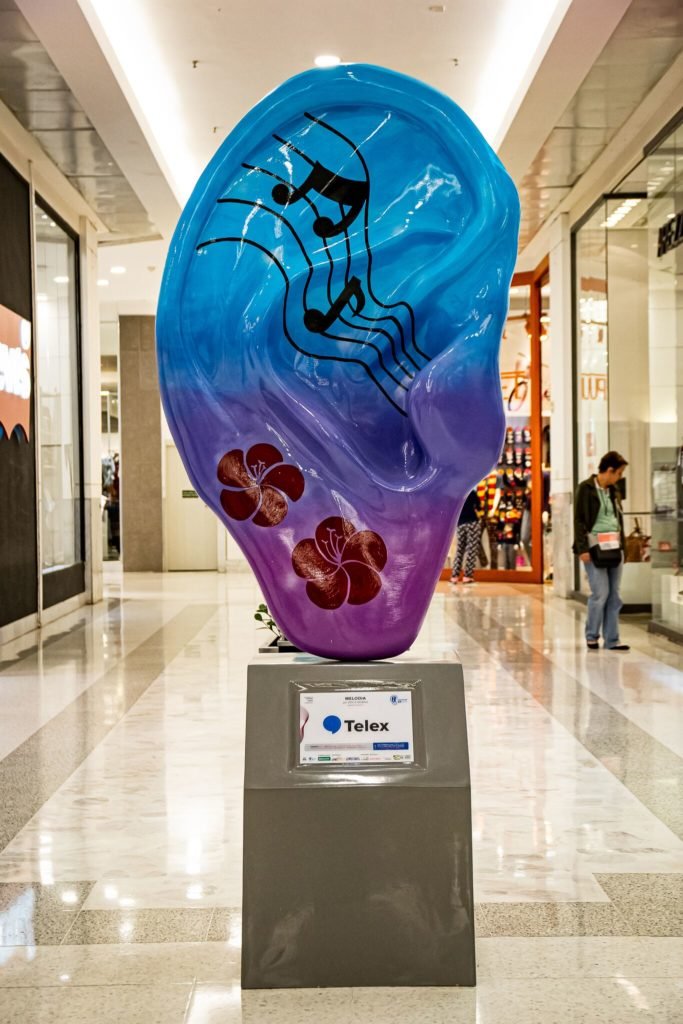 Descrição da imagem #PraCegoVer: Imagem de uma escultura de orelha gigante. A peça é azul e está estilizada com imagens que remetem à notas musicais. Fim da descrição.