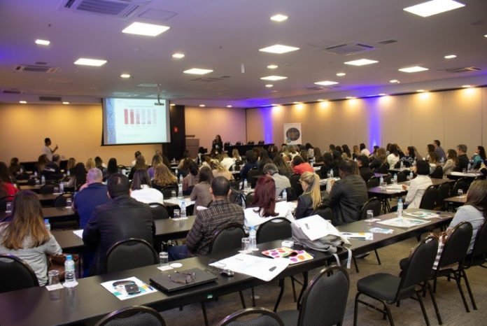 Descrição da imagem #PraCegoVer: Auditório do evento sobre atrofia muscular espinhal em uma edição anterior. No auditório, várias pessoas estão sentadas, acompanhando uma apresentação que está sendo feita em um telão. Fim da descrição.