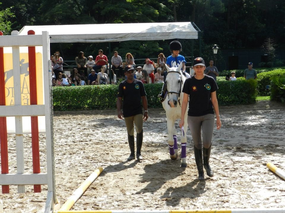 Descrição da imagem: foto de dois profissionais de equoterapia no setting terapêutico. Eles estão conduzindo um cavalo branco durante o atendimento de uma criança. Fim da descrição.