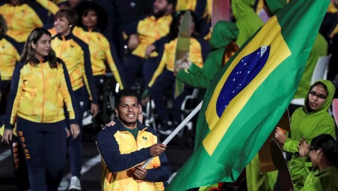 Descrição da imagem #PraCegoVer: O paratleta Leomon Moreno como porta-bandeira na cerimônia de abertura dos Jogos Parapan-Americanos de Lima. Ele está segurando a bandeira brasileira e usa um agasalho amarelo com manga azul escuro. Leomon está sorrindo. Atrás dele, vemos outros atletas brasileiros. Fim da descrição.