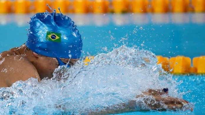 Descrição da imagem #PraCegoVer: Atleta da natação está dentro da piscina, durante disputa em Lima. Na imagem, vemos o atleta de perfil, e ele está usando toca azul com a bandeira do Brasil. Vemos detalhes do movimento da água. Fim da descrição.