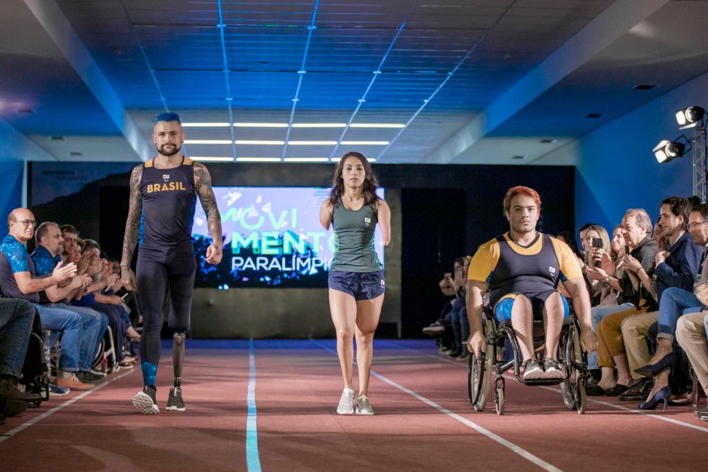 Descrição da imagem #PraCegoVer. Três atletas desfilam durante lançamento dos uniformes do Parapan em Lima. Da esquerda para a direita, temos o atleta Vinicius Rodrigues, que usa uma prótese na perna esquerda. Ele é moreno, tem pele clara e usa uma regata azul marinho com o nome Brasil, em amarelo, e uma bermuda azul marinho. No meio está a atleta Dayane Silva, que tem uma malformação nos braços. Ela é morena, tem a pele clara, e está usando uma regata, verde escuro e um shorts azul marinho. O terceiro é o atleta Matheus Assis, que é cadeirante. Ele é moreno claro e está usando um uniforme azul marinho e amarelo. O desfile foi realizado na pista de atletismo do CT Paralímpico, por isso, o piso é marrom com listras divisórias brancas. Fim da descrição.