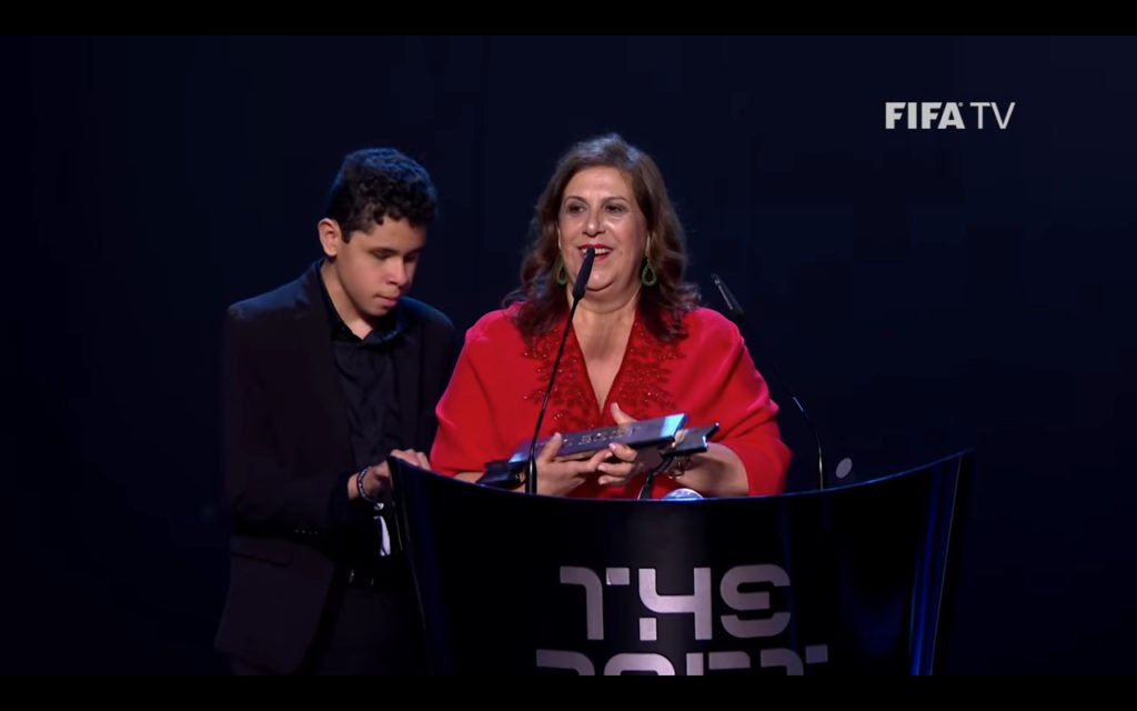 Descrição da imagem #PraCegoVer: Silvia e Nickollas durante o discurso após receberem o prêmio Fifa Fan Award. Eles estão no palco do evento, atrás do púlpito, que é preto. O fundo do palco também é todo preto. Do lado esquerdo está Nickollas, um menino de 12 anos, moreno com pele clara e cabelos curtos e pretos. Ele está vestindo um terno preto. Do lado direito está Silvia, uma mulher morena, de pele clara e cabelos compridos e castanhos. Ela está usando um vestido vermelho e segura o troféu. Fim da descrição