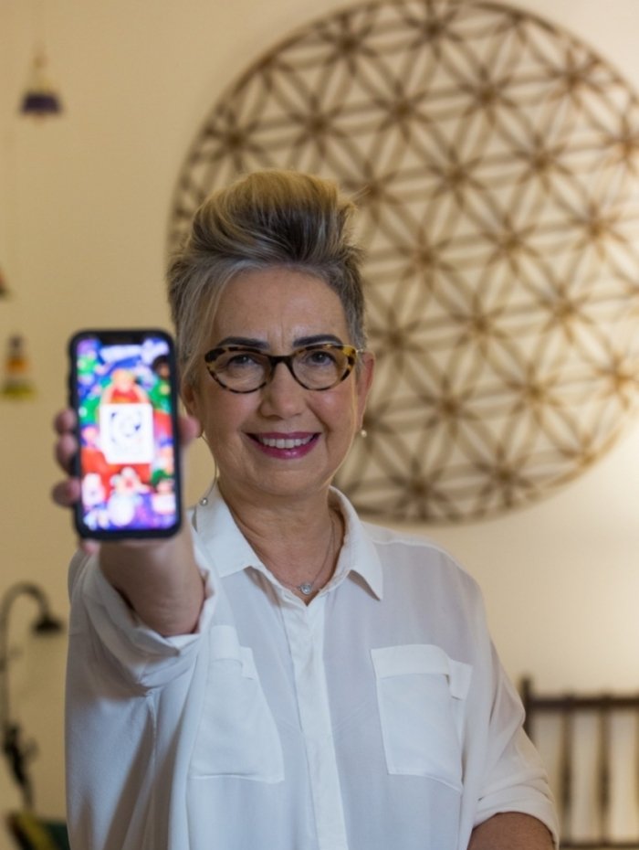 Descrição da imagem #PraCegoVer: Claudia Werneck está segurando um celular com o app Vem Ca aberto. Claudia é uma mulher loira, com cabelos curtos. Ela tem pele clara, usa óculos e uma camisa branca. Claudia está sorrindo. Fim da descrição.