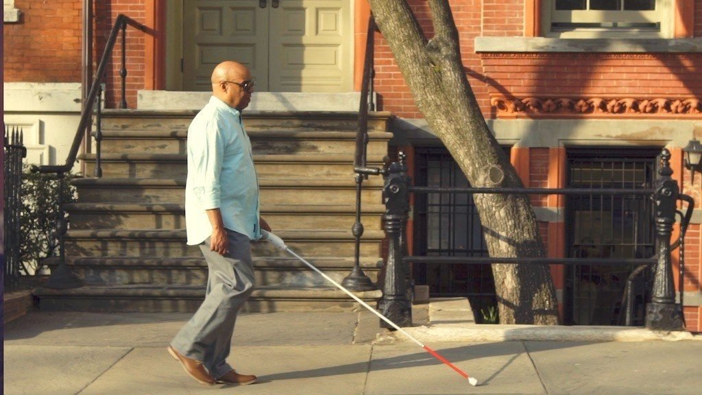 Descrição da imagem #PraCegoVer: Um homem caminha pela rua. Ela segura o dispositivo WeWalk acoplado a uma bengala. Fim da descrição.