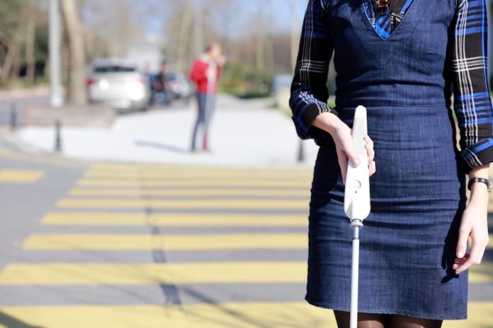 Descrição da imagem #PraCegoVer: Uma mulher caminha pela rua. Ela segura o dispositivo WeWalk acoplado a uma bengala. Fim da descrição.