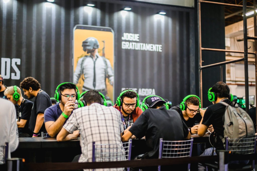 Descrição da imagem #PraCegoVer: Vários rapazes estão sentados em uma mesa, jogando videgame durante a última edição do Brasil Game Show. Eles estão usando fones de ouvido verdes. Fim da descrição.