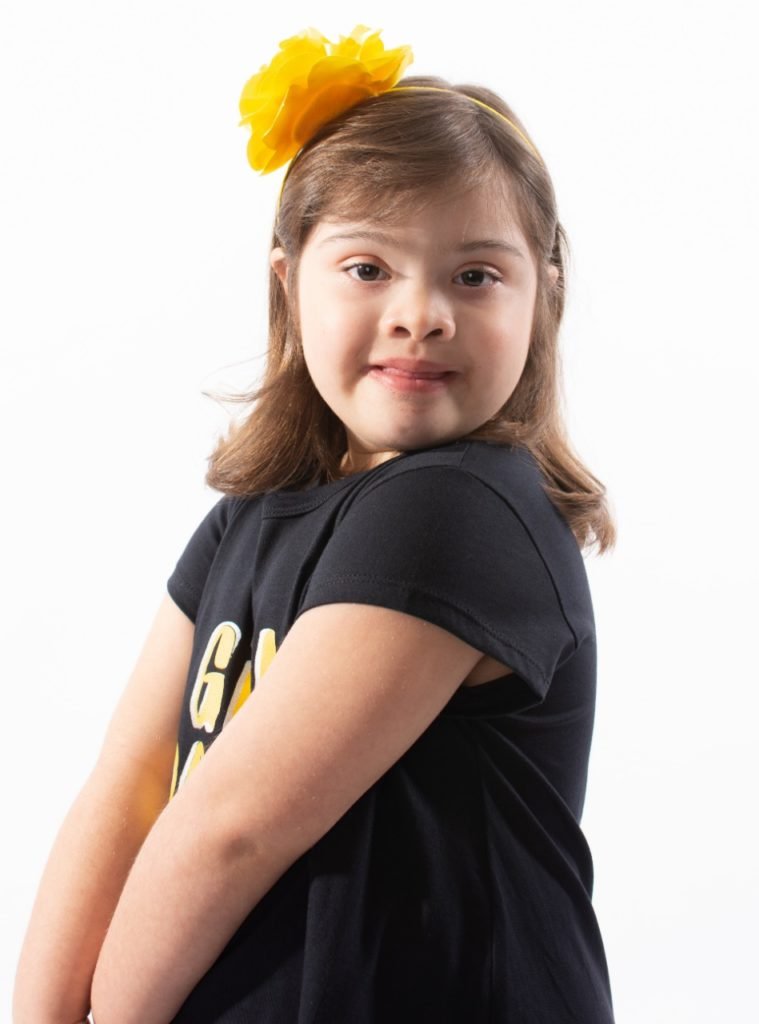 Descrição da imagem #PraCegoVer: A menina Helena Nistal está posando para a foto. Ela é uma menina morena, de pele clara e cabelos pretos, lisos, na altura dos ombros. Helena está com o tronco virado de lado e usa uma camiseta preta e uma tiara amarela. Fim da descrição.