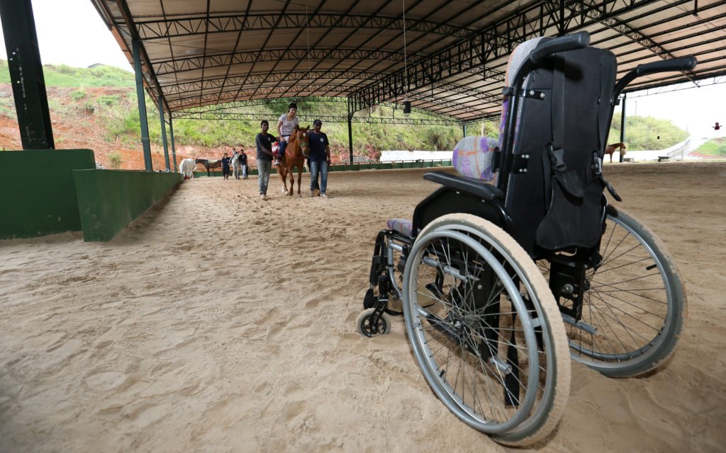 Descrição da imagem #PraCegoVer: Picadeiro da Hípica Santa Terezinha. Vemos, em primeiro plano, uma cadeira de rodas, e ao fundo, as pessoas durante a sessão no cavalo. Fim da descrição.