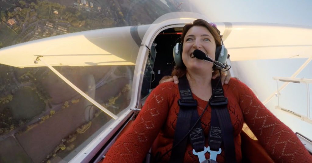 Descrição da imagem #PraCegoVer: Cena do filme 'Fecha os olhos e voa'. Uma mulher cega está na cabine de um avião monomotor. Ela está sentindo os movimentos da aeronave e sorri. Fim da descrição
