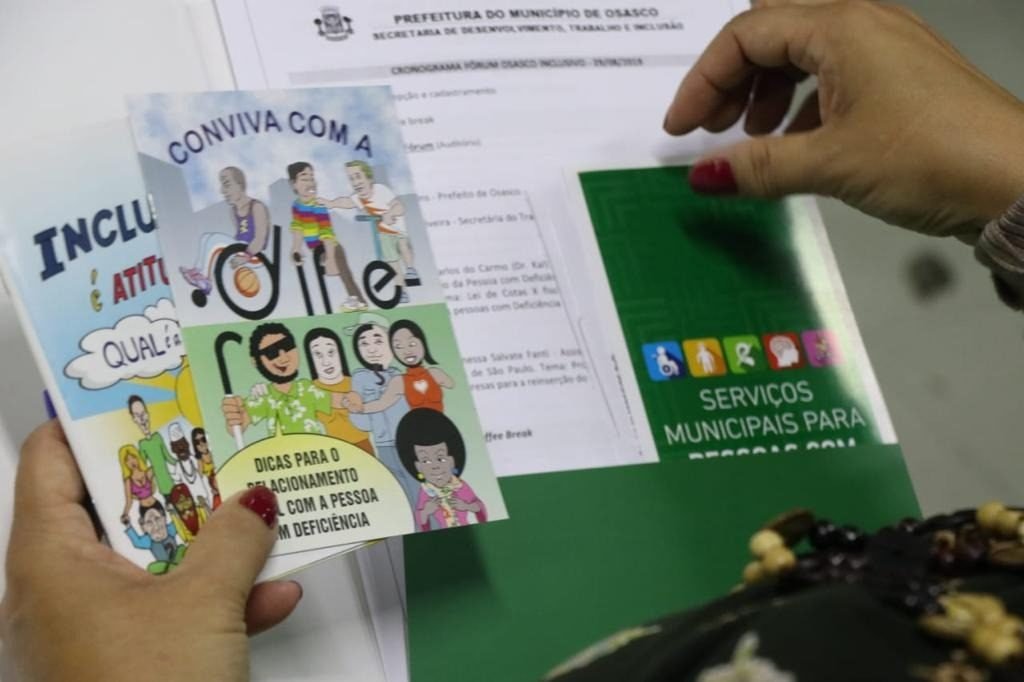 Descrição da imagem #PraCegoVer: Uma mulher segura, na mão esquerda, cartilhas sobre empregabilidade para pessoas com deficiência. Na mão direita, ela segura panfletos. Fim da descrição.