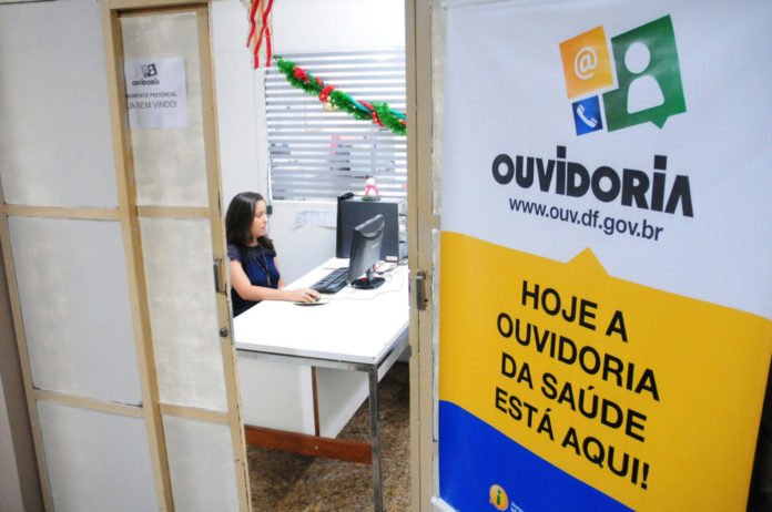 Descrição da imagem #PraCegoVer: Entrada da ouvidoria da saúde no Distrito Federal. Dentro da sala est[a uma mulher sentada, usando um computador. Fim da descrição.