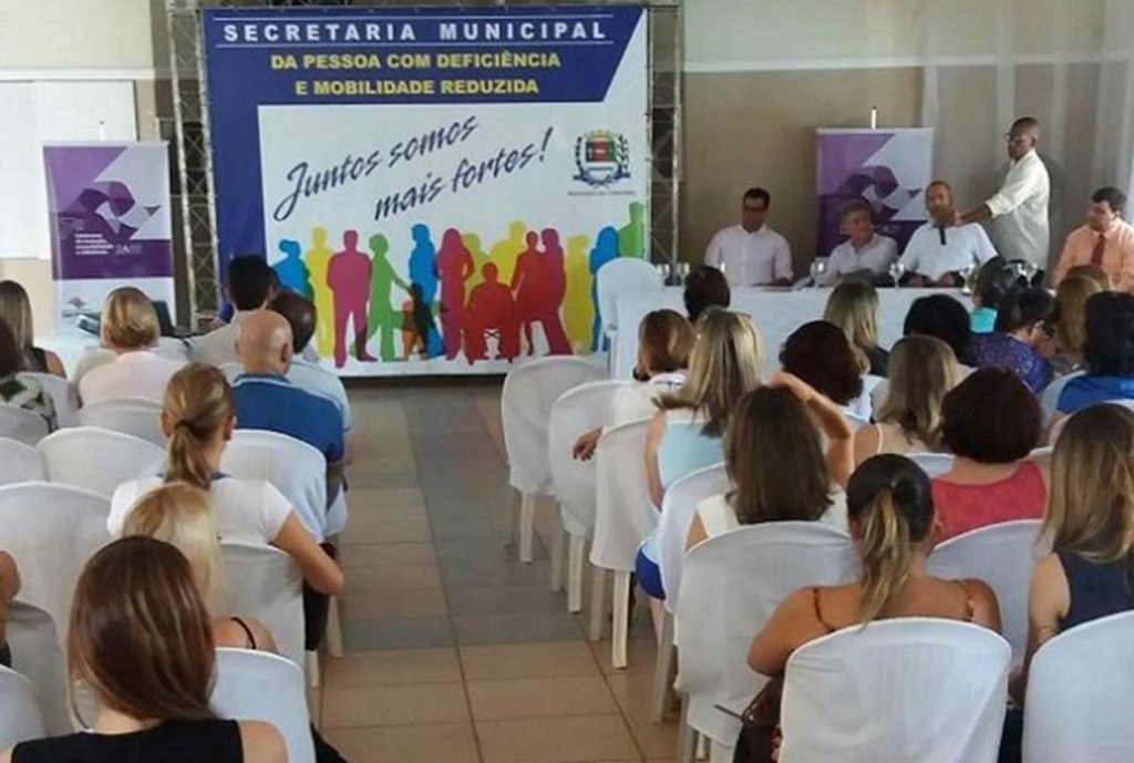 Descrição da imagem #PraCegoVer: Uma sala onde está sendo realizada uma das edições da Caravana da Inclusão. Várias pessoas estão sentadas em cadeiras brancas e acompanham um discurso. Algumas autoridades estão sentadas em uma mesa, ao lado de um banner grande e colorido, que traz as informações do evento. Fim da descrição.