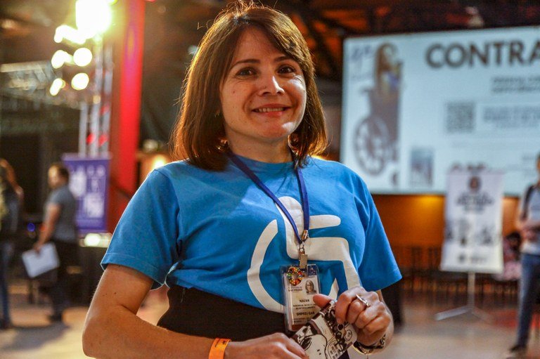 Descrição da imagem #PraCegoVer: Uma mulher está sorrindo. Ela é morena e tem cabelos castanhos, lisos, na altura dos ombros. Ela está usando uma camiseta azul com uma imagem branca do símbolo da deficiência física. Fim da descrição.
