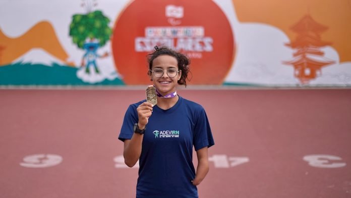 Descrição da imagem #PraCegoVer: A paratleta Maria Clara Augusto da Silva está na pista de atletismo, segurando a medalha de ouro. Ela é uma menina morena, de pele clara, cabelos castanhos presos em um rabo de cavalo e usa óculos. Ela tem uma malformação no braço esquerdo. Fim da descrição.