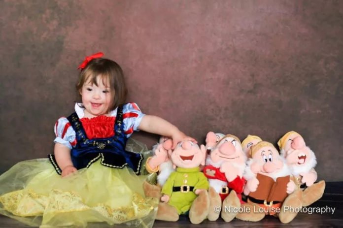 Descrição da imagem: foto de reprodução do filme ‘Branca de Neve e os Sete Anões’. Uma menina com síndrome de Down está vestida de Branca de Neve, protagonista do filme. Ela está sentada ao lado de ursinhos representando os Sete Anões. Fim da descrição.