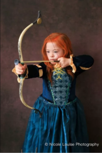 Descrição da imagem: foto de reprodução do filme ‘Valente’. Uma menina com síndrome de Down está vestida de Valente, protagonista do filme. Ela está usando um vestido azul escuro e faz pose com um arco e flecha. Fim da descrição.