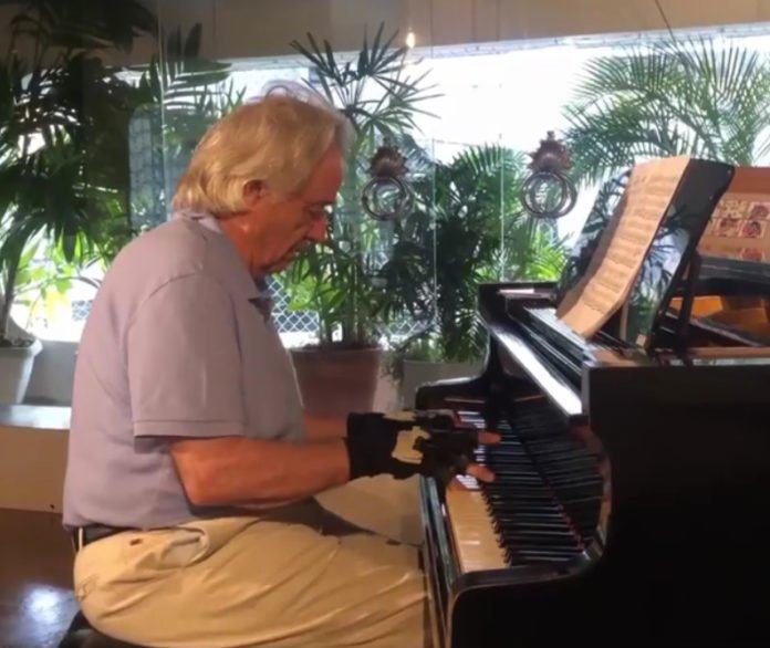 Descrição da imagem #PraCegoVer: O maestro João Carlos Martins sentado à frente de seu piano. Ele está usando as luvas biônicas que lhe permitiram voltar a tocar o instrumento. João é um senhor de cabelos brancos. Ele veste uma calça bege e uma camisa polo azul. Ao fundo vemos folhagens verdes de um jardim. Fim da descrição.